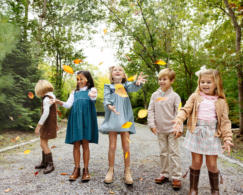 Five children stand on a gravel path in a wooded area, smiling and tossing autumn leaves into the air. They are dressed for fall in boots, sweaters, and the Long Sleeve Daphne Dress - Grier Green Herringbone by The Beaufort Bonnet Company. Behind them, green and yellow trees create a vibrant backdrop.