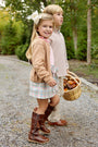 Two young children walk along a gravel path, the girl smiling at the camera in The Beaufort Bonnet Company’s Beasley Bow Skirt – Manalapan Plaid paired with boots, while the boy carries a basket of autumn leaves. They are surrounded by trees and greenery.