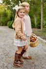 Two young children walk along a gravel path, the girl smiling at the camera in The Beaufort Bonnet Company’s Beasley Bow Skirt – Manalapan Plaid paired with boots, while the boy carries a basket of autumn leaves. They are surrounded by trees and greenery.