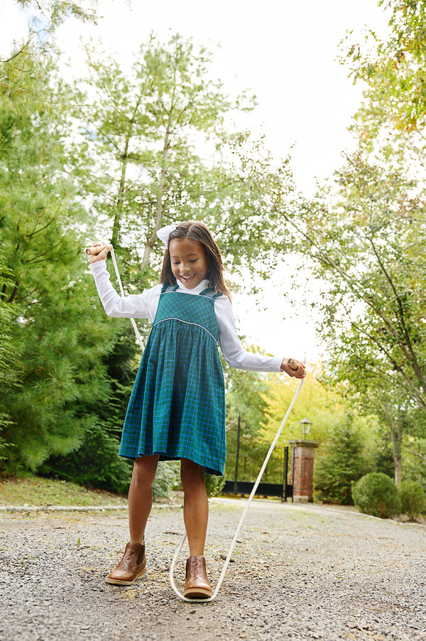A young girl wearing The Beaufort Bonnet Company’s Demi Dress in Woodlake Point Plaid with Worth Avenue White and brown boots smiles as she jumps rope on a gravel path, surrounded by green trees on a sunny day.
