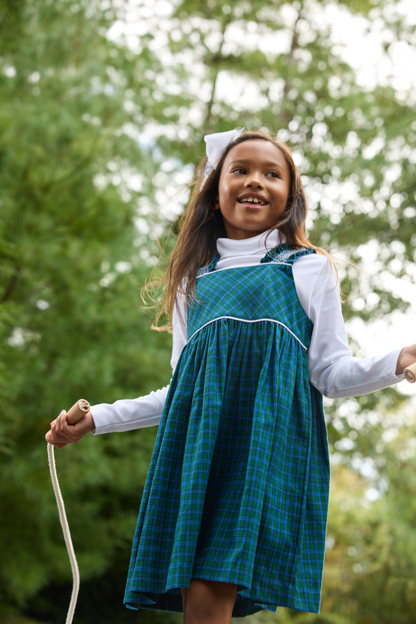 A young girl wearing The Beaufort Bonnet Company’s Demi Dress in Woodlake Point Plaid with Worth Avenue White and a white hair bow smiles as she jumps rope outdoors, surrounded by lush green trees.