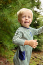 A cheerful blonde child poses outdoors by green pine trees dressed in The Beaufort Bonnet Company’s Rayburn Romper – Grier Green Stripe with Nantucket Navy, crafted from soft Pima cotton for a cozy and comfortable fall outfit.