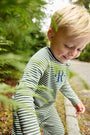 A young child smiles and walks outdoors near lush green foliage and a gravel path, dressed in The Beaufort Bonnet Company’s Rayburn Romper in Grier Green Stripe with Nantucket Navy—a cozy and adorable fall outfit.