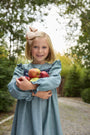 A young girl with blonde hair and a large bow is smiling as she stands on a gravel path surrounded by green trees, holding several apples and wearing The Beaufort Bonnet Company’s Long Sleeve Daphne Dress in Grier Green Herringbone.