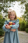 A young girl with blonde hair and a large bow is smiling as she stands on a gravel path surrounded by green trees, holding several apples and wearing The Beaufort Bonnet Company’s Long Sleeve Daphne Dress in Grier Green Herringbone.