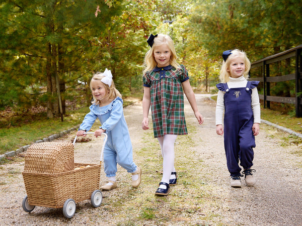 Three young girls walk along a gravel path in a park, smiling and enjoying themselves. One girl pushes a wicker pram, and all wear colorful outfits from The Beaufort Bonnet Company—including Violet’s Vintage Overalls in Nantucket Navy with Worth Avenue White—with bows adorning their hair. Lush green trees line the path as they explore together.