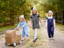Three young girls walk along a gravel path in a park, smiling and enjoying themselves. One girl pushes a wicker pram, and all wear colorful outfits from The Beaufort Bonnet Company—including Violet’s Vintage Overalls in Nantucket Navy with Worth Avenue White—with bows adorning their hair. Lush green trees line the path as they explore together.