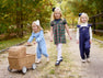 Three young girls walk outside on a gravel path; one pushes a wicker stroller. Dressed in blue and plaid outfits, including the Tatum’s Turtleneck & Onesie in Nantucket Navy Microdot from The Beaufort Bonnet Company, they are surrounded by green trees.