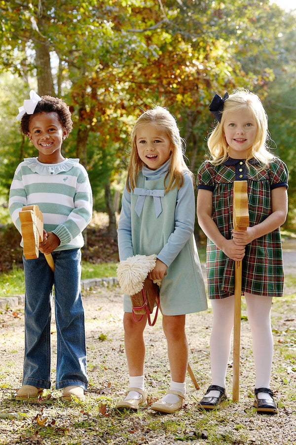 Three young girls stand outdoors, beaming as they hold toy hobby horses. They wear vibrant outfits from The Beaufort Bonnet Company, including one girl dressed in the Jilly Jumper in Old Greenwich Green Corduroy, perfectly paired with a Sarasota Sky Bow in her hair. Sunlight streams through the trees behind them.