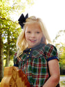 A young girl with long blonde hair and a black bow wears The Beaufort Bonnet Company’s Dale Dress - Pinehill Plaid with Nantucket Navy and smiles outdoors beside a wooden rocking horse, surrounded by sunlit trees in the background.
