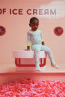 A young child wearing The Beaufort Bonnet Company’s Fallon Footie Pajamas - Ice Cream Daydream (Blueberry Blue) with Nana Banana sits on a bench above a pit filled with pink balloon shapes, with “OF ICE CREAM” visible on a pink tiled wall behind them.