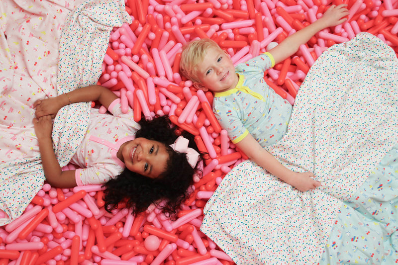 Two children—one with curly dark hair and the other with short blond hair—smile while lying on their backs amid a sea of pink balloon shapes, cozily wrapped in The Beaufort Bonnet Company’s Silent Night Throw - Ice Cream Daydream (Blueberry Blue). Their patterned blankets feature colorful designs, and both appear happy and relaxed.