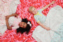 Two children—one with curly dark hair and the other with short blond hair—smile while lying on their backs amid a sea of pink balloon shapes, cozily wrapped in The Beaufort Bonnet Company’s Silent Night Throw - Ice Cream Daydream (Blueberry Blue). Their patterned blankets feature colorful designs, and both appear happy and relaxed.