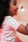 A young child with curly hair smiles while wearing The Beaufort Bonnet Company’s Payton Pajama Shorts Set in Ice Cream Daydream (Strawberry Pink), standing against a soft pink background.