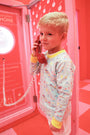 A young boy with blonde hair holds a red phone receiver to his ear in a pink room, standing next to a pink public payphone. He’s wearing The Beaufort Bonnet Company’s Cassidy Comfy Crewneck - Ice Cream Daydream (Blueberry Blue), featuring playful colorful prints—a perfect layering piece crafted from premium materials.
