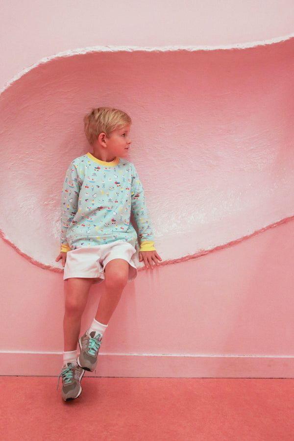 A young boy wearing The Beaufort Bonnet Company’s Cassidy Comfy Crewneck in Ice Cream Daydream (Blueberry Blue) and white shorts sits against a pink wall with a curved, textured indentation, looking to his left.