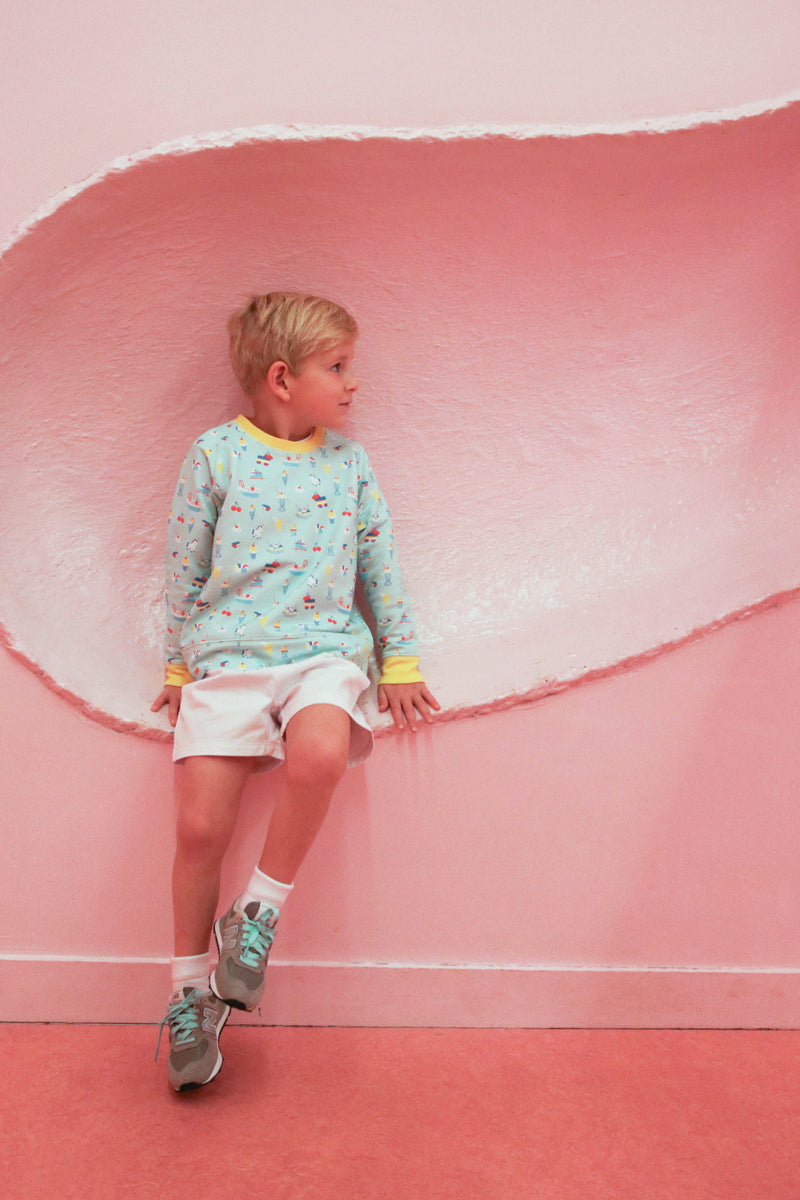 A young boy wearing The Beaufort Bonnet Company’s Cassidy Comfy Crewneck in Ice Cream Daydream (Blueberry Blue) and white shorts sits against a pink wall with a curved, textured indentation, looking to his left.