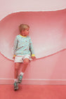 A young boy wearing The Beaufort Bonnet Company’s Cassidy Comfy Crewneck in Ice Cream Daydream (Blueberry Blue) and white shorts sits against a pink wall with a curved, textured indentation, looking to his left.