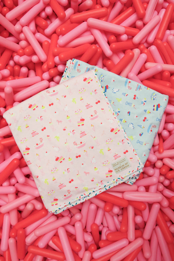 Two patterned baby blankets—one pink and one blue—are laid out on a background of numerous pink and red balloon-like shapes, alongside The Beaufort Bonnet Company’s Silent Night Throw - Ice Cream Daydream (Blueberry Blue).