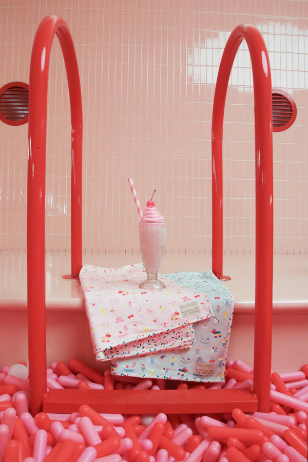 A pink and white milkshake with a cherry and straw sits atop pastel towels, including The Beaufort Bonnet Company’s Silent Night Throw in the Ice Cream Daydream (Blueberry Blue) print, draped over a red pool ladder. The scene is surrounded by pink and red balloon-like objects in a tiled room.