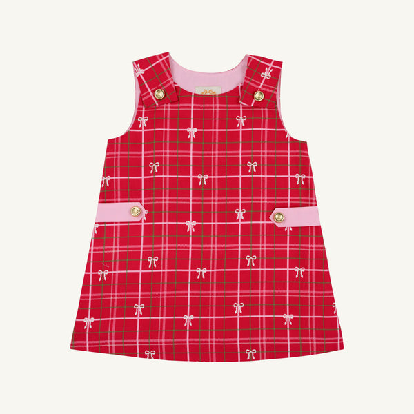 The Beaufort Bonnet Company’s Janie Jumper in Torrington Tartan with Pier Party Pink is a girls’ flannel dress for toddlers, featuring a sleeveless red tartan design, accented with small white bows, pink side bands, and gold buttons on the shoulders and waist. The dress is shown against a plain white background.