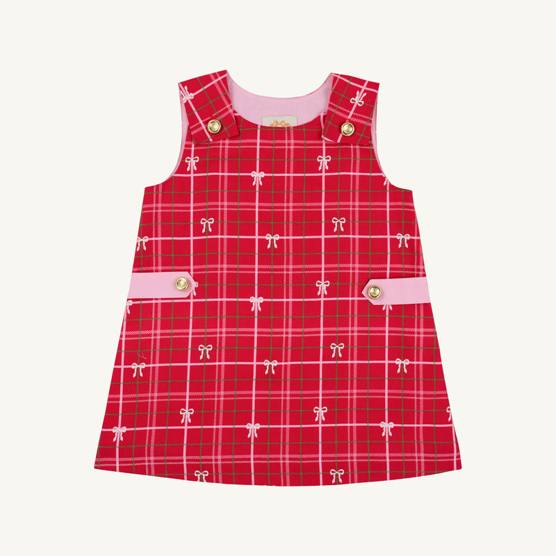 The Beaufort Bonnet Company’s Janie Jumper in Torrington Tartan with Pier Party Pink is a girls’ flannel dress for toddlers, featuring a sleeveless red tartan design, accented with small white bows, pink side bands, and gold buttons on the shoulders and waist. The dress is shown against a plain white background.