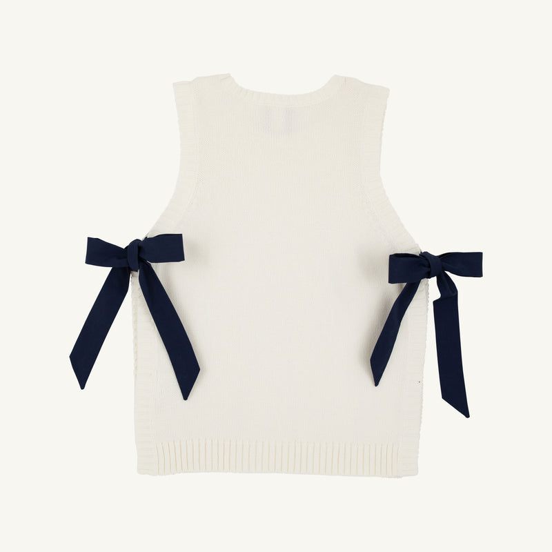 Introducing the Shae Sweater Vest - Palmetto Pearl with Nantucket Navy by The Beaufort Bonnet Company: a sleeveless white knit vest with ribbed edges, adorned with two navy blue ribbon bows tied at the sides. Made from 100% cotton, this sweater is a charming addition to any girl’s autumn wardrobe.