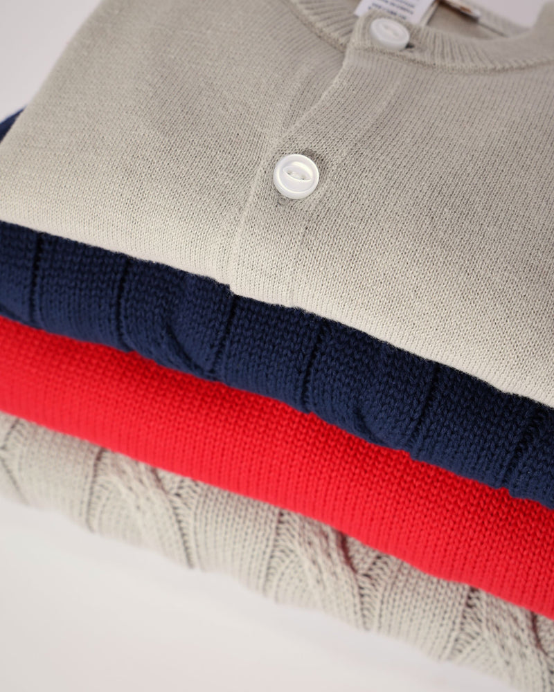 A stack of Cambridge Cardigan - Grantley Gray, navy blue, and bright red cardigans folded for a little one’s wardrobe.