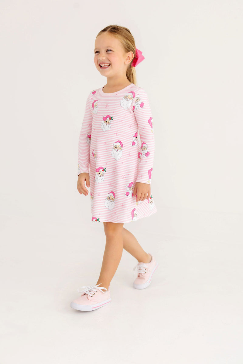 A smiling girl wears a Long Sleeve Polly Play Dress - Dear Santa (Palm Beach Pink Stripe) with pink striped Santa Claus graphics and light pink sneakers.