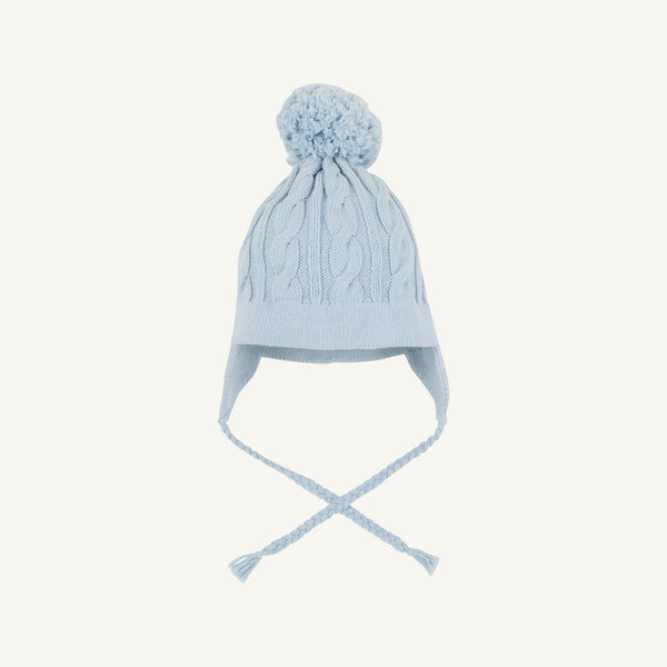 The Beaufort Bonnet Company’s Parrish Pom Pom Hat in Buckhead Blue is a light blue cotton knit hat featuring a classic cable knit pattern, ear flaps, braided tassels, and a large pom-pom on top. It is displayed on a plain white background.