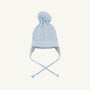 The Beaufort Bonnet Company’s Parrish Pom Pom Hat in Buckhead Blue is a light blue cotton knit hat featuring a classic cable knit pattern, ear flaps, braided tassels, and a large pom-pom on top. It is displayed on a plain white background.