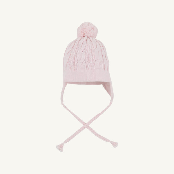 The Parrish Pom Pom Hat - Palm Beach Pink by The Beaufort Bonnet Company is a light pink cotton knit hat showcasing a classic cable-knit pattern, playful pom-pom on top, ear flaps on each side, and two braided tassels, all presented against a plain white background.