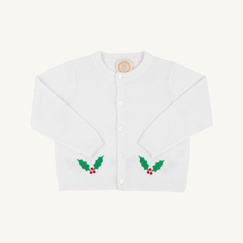 The Beaufort Bonnet Company’s Cambridge Cardigan in Worth Avenue White features long sleeves, front buttons, and festive green holly leaf with red berry designs near the bottom on each side. This cheerful baby sweater stands out against its plain light background, making it a perfect choice for the holiday season.