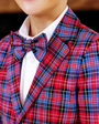 A person wearing a Bucktown Tartan plaid suit jacket paired with The Beaufort Bonnet Company’s Baylor Bowtie in Bucktown Tartan and a white dress shirt, smiling slightly.