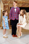 Three children dressed in formal clothes smile and interact in a living room. A boy stands wearing The Beaufort Bonnet Company’s Prep School Pants - Newport Night Corduroy with Richmond Red Stork, while two girls, one in blue and one in cream, sit and stand nearby—all appearing cheerful.