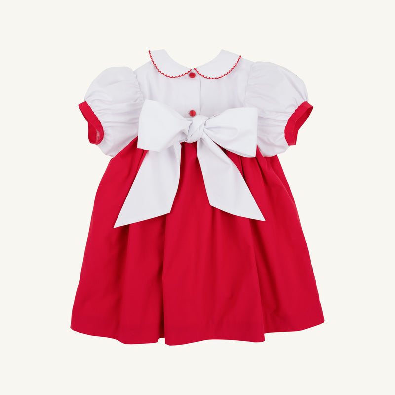 The Mary Dal Dress - Richmond Red & Worth Avenue White with Christmas Tree by The Beaufort Bonnet Company is a festive holiday dress for babies, featuring puffy short sleeves, a Peter Pan collar, charming red buttons, and a large white bow tied at the back—perfect for special holiday occasions or as a Christmas tree dress.