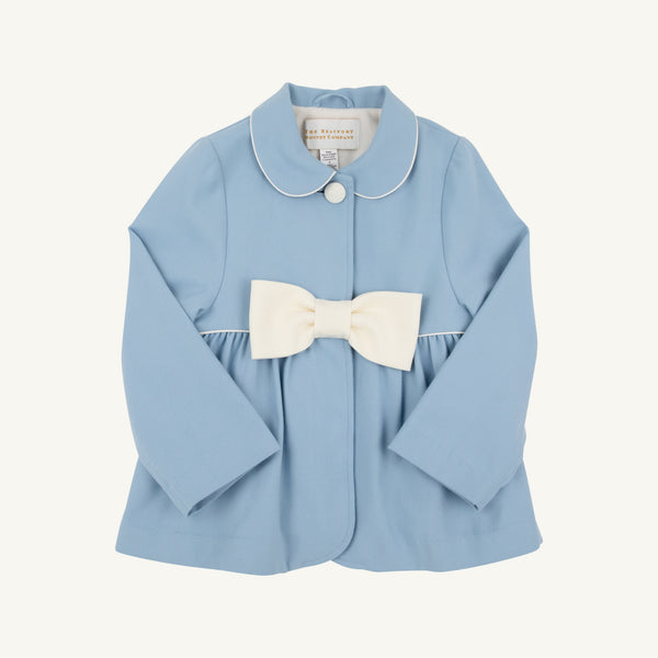 A light blue Pippa Peacoat by The Beaufort Bonnet Company in Barrington Blue with Palmetto Pearl, designed for toddlers and featuring long sleeves, a white Peter Pan collar trim, a single front button, and a large white bow at the waist.