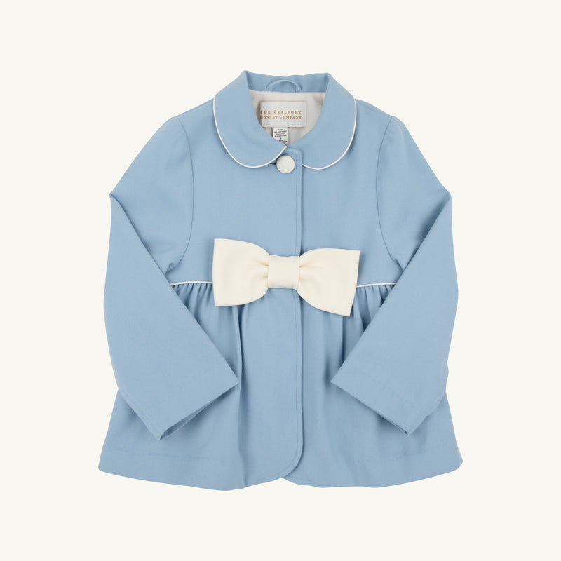 A light blue Pippa Peacoat by The Beaufort Bonnet Company in Barrington Blue with Palmetto Pearl, designed for toddlers and featuring long sleeves, a white Peter Pan collar trim, a single front button, and a large white bow at the waist.