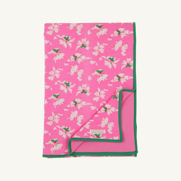 A folded Silent Night Throw - Bows of Holly with Kiawah Kelly Green by The Beaufort Bonnet Company, featuring a pink Pima Cotton Knit decorated with a white holly floral pattern and accented by green trim. The blanket is partially flipped to reveal its plain pink underside and a small fabric label. Perfect as a festive holiday throw blanket.