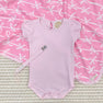 A pale pink baby bodysuit, a Parker Paci Clip - Pink Greenbrier Gingham with Palm Beach Pink, and a pink blanket with a white bow pattern.