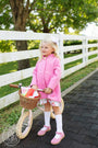 A young blonde girl wearing The Beaufort Bonnet Company’s Penelope Peacoat in Hamptons Hot Pink with Richmond Red, a white patterned dress, and white knee-high socks stands next to a pink balance bike with a wicker basket filled with wrapped gifts. She has a white bow in her hair and is standing by a white fence.