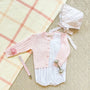 A pink cable-knit cardigan, white bubble romper, matching floral bonnet, silver rattle, and a Parker Paci Clip - Pink Greenbrier Gingham with Palm Beach Pink pacifier, all arranged on a plaid blanket.