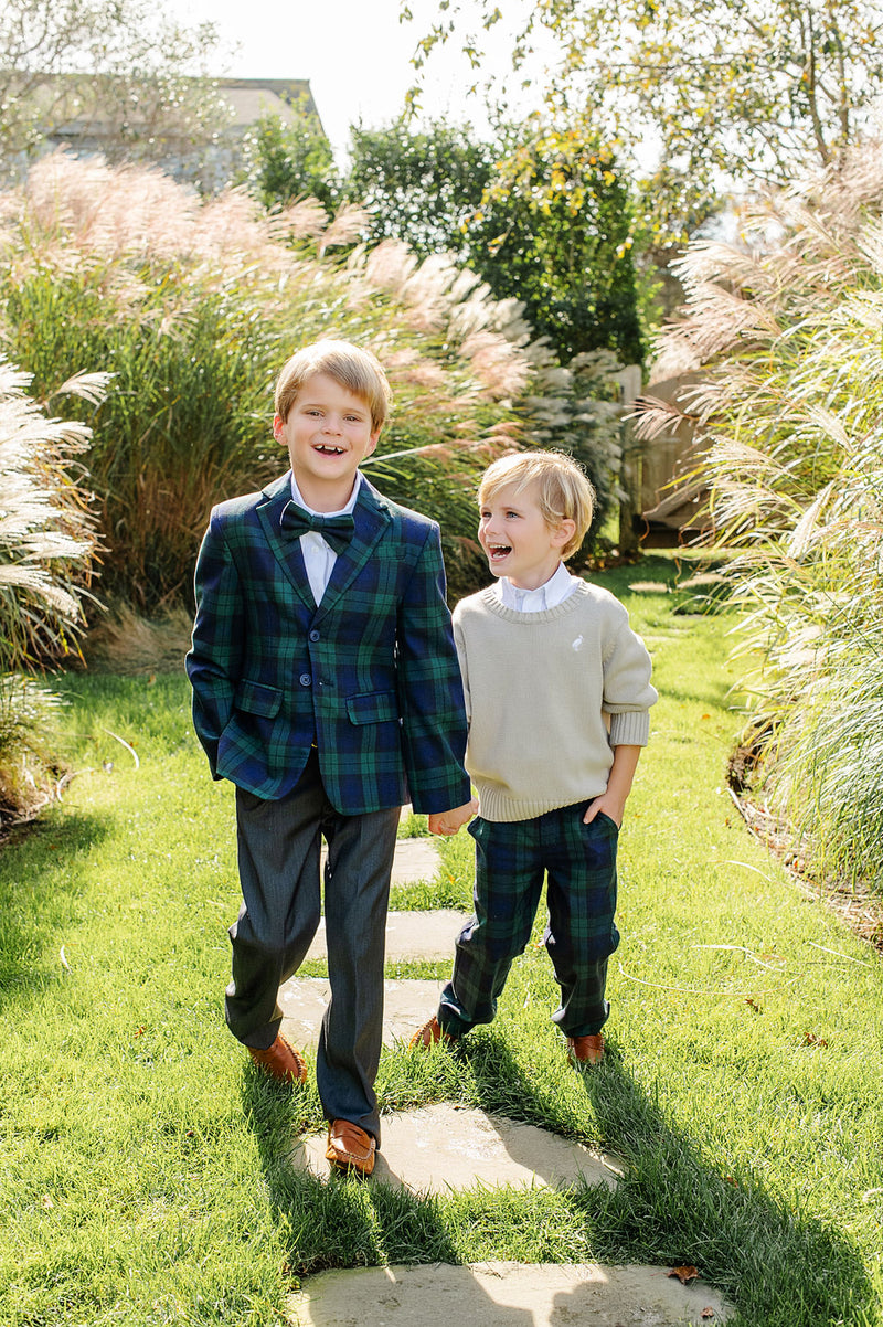 Two young boys walk along a garden path, smiling. The older boy wears a plaid suit paired with The Beaufort Bonnet Company’s Baylor Bowtie in Berwick Black Watch, while the younger boy sports a beige sweater and plaid pants. Sunlight filters through tall grasses and trees around them.