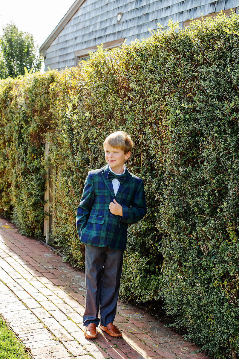 A young boy in a plaid blazer, dress shirt, gray pants, and a Baylor Bowtie – Berwick Black Watch by The Beaufort Bonnet Company stands on a brick path beside a tall green hedge, with one hand in his pocket, looking to the side and smiling.
